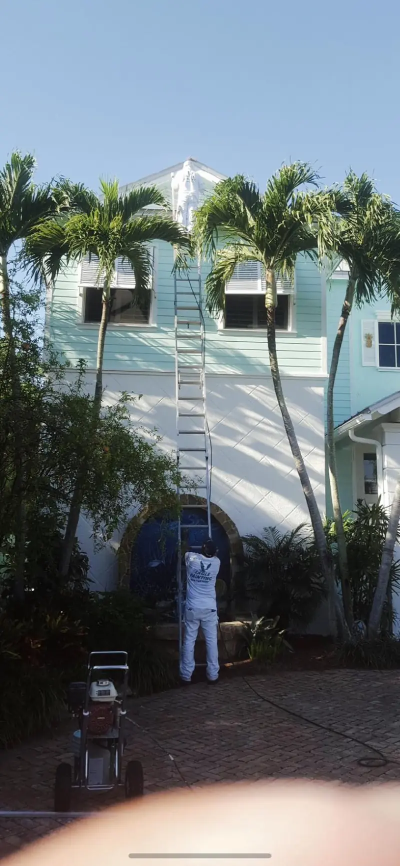 Painter on an extension ladder repainting a two-story house with palm trees
